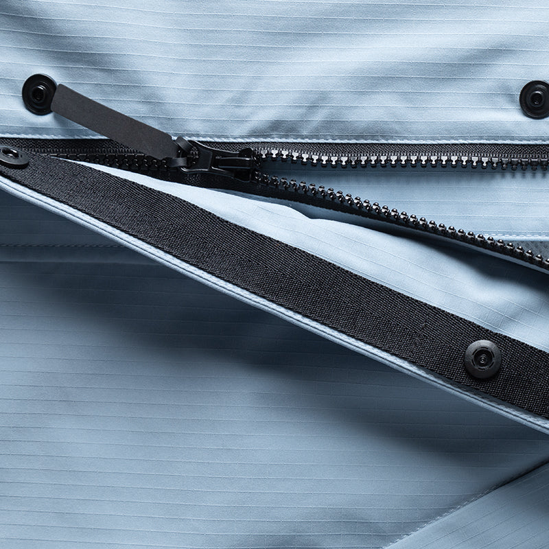 Nahaufnahme einer hellblauen Jacke mit Reißverschluss, schwarzem Streifen und Druckknöpfen auf einem strukturierten Stoff.