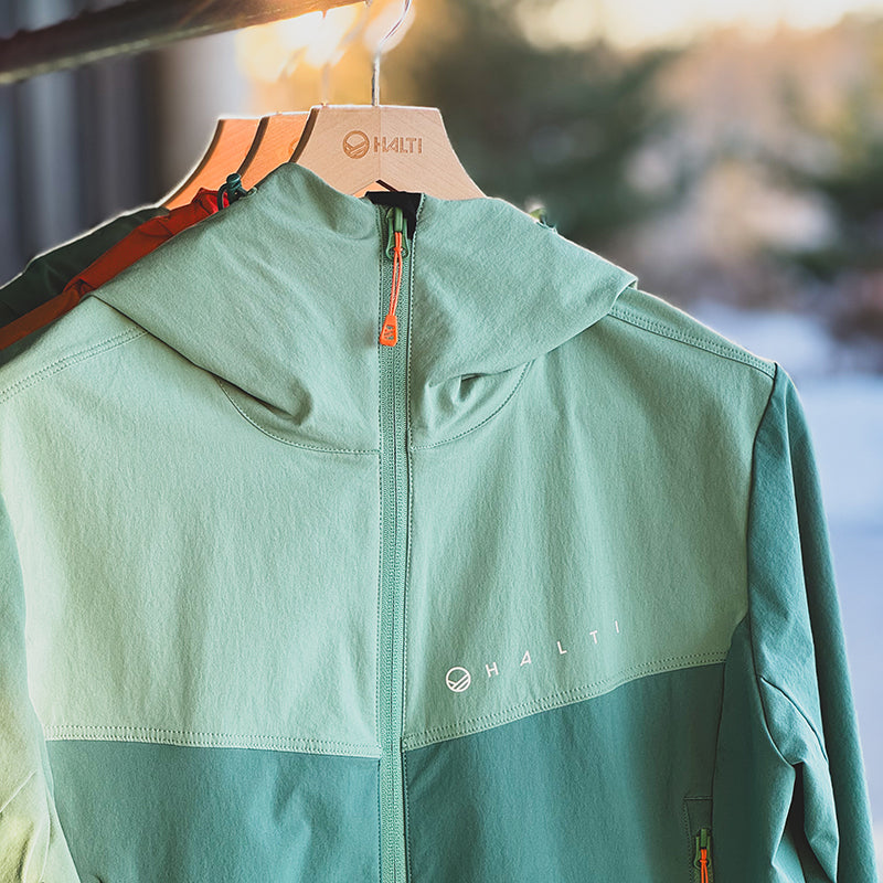 Hellgrüne Halti Pallas Evo X-Stretch-Jacke Plus Damen mit Kapuze, im Freien auf Holzbügeln hängend.