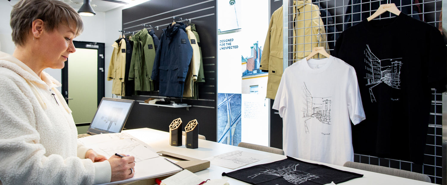 Eine Person skizziert an einem Schreibtisch mit ausgestellten Kleidungsstücken, Hemden und Auszeichnungen in einer modernen Studioumgebung.