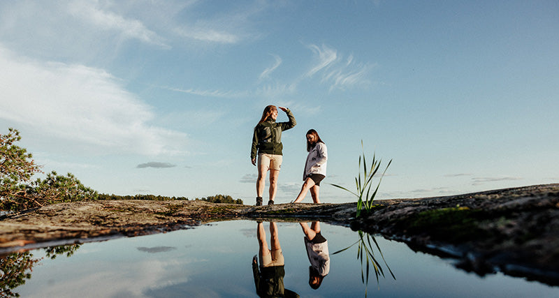 Zwei Personen stehen auf einem Felsen in der Nähe eines kleinen Wasserbeckens, in dem sie sich spiegeln, während über ihnen ein blauer Himmel zu sehen ist.