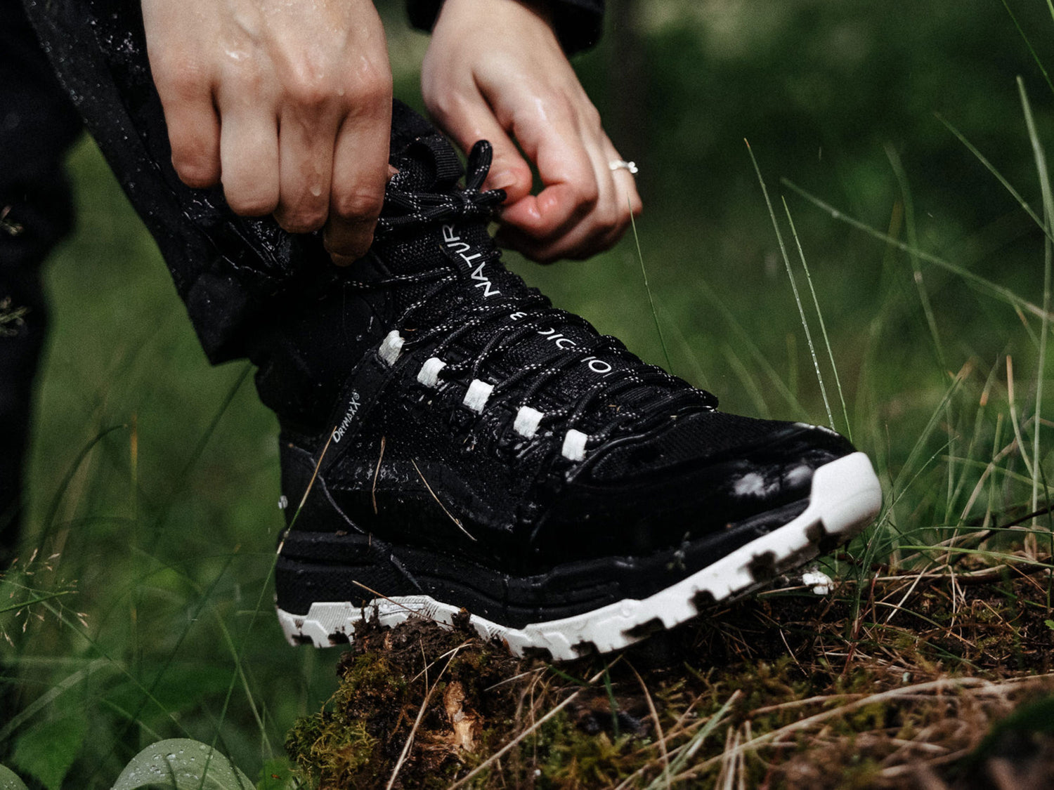 Person, die die Schnürsenkel eines schwarzen Wanderschuhs mit weißer Sohle bindet, während sie im Freien auf Gras und Moos steht.