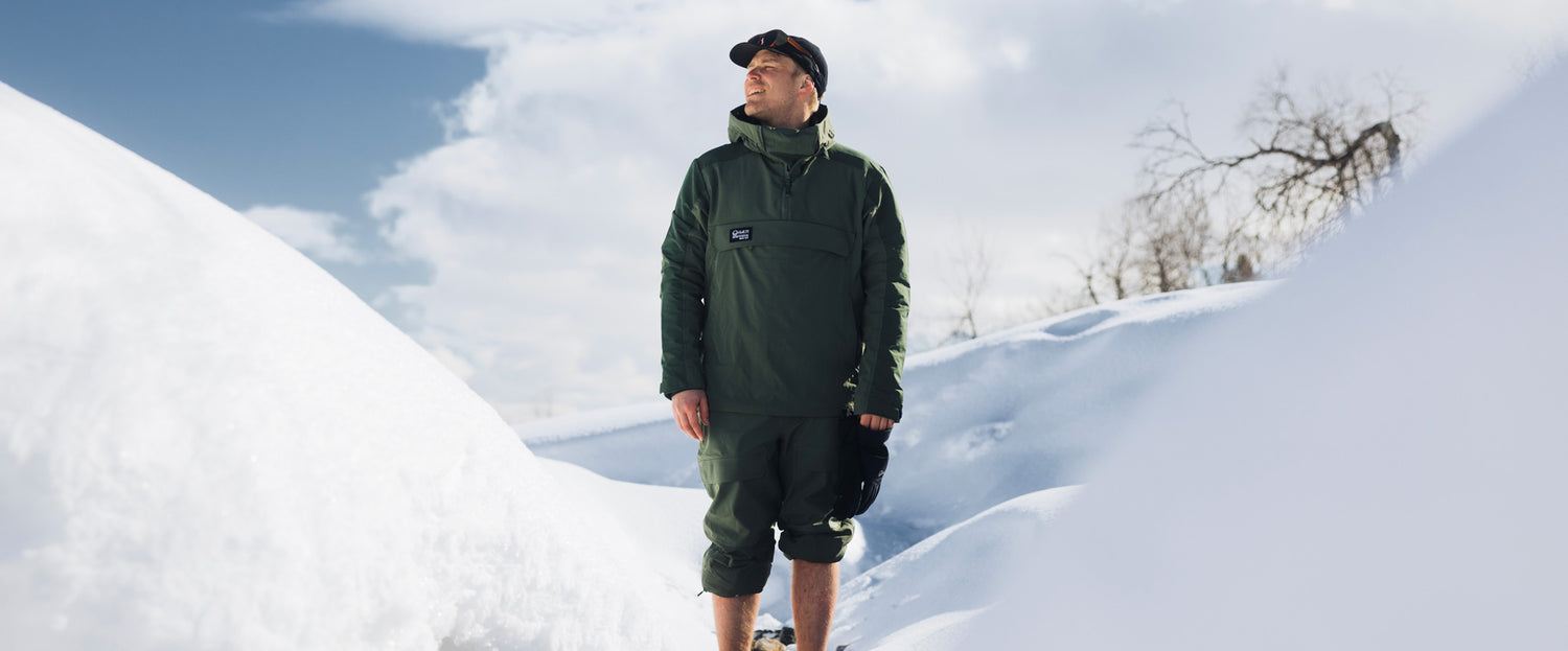Eine Person mit grüner Jacke und Hut steht im tiefen Schnee und blickt nach oben, während der Himmel bewölkt ist und die Bäume im Hintergrund kahl sind.