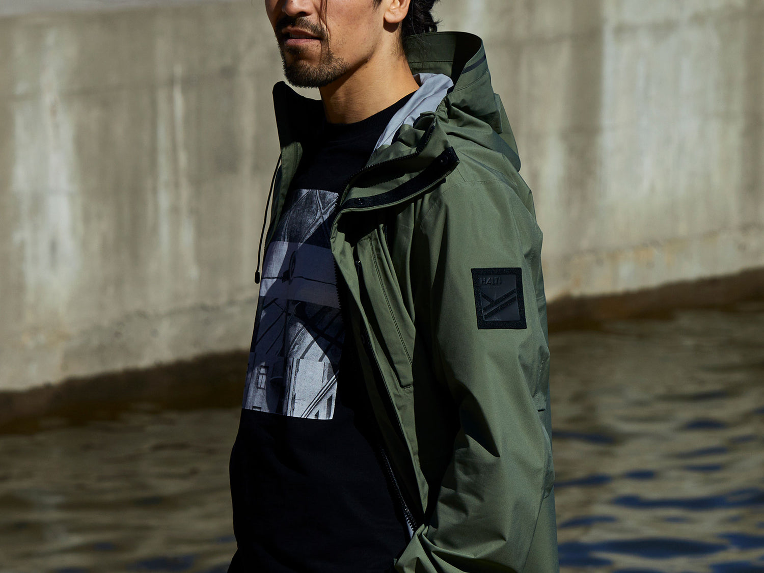 Eine Person mit grüner Jacke und schwarzem T-Shirt steht in der Nähe einer Betonwand und Wasser.