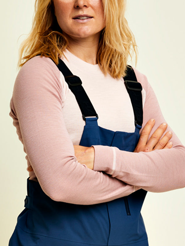 Eine Person mit blondem Haar verschränkt die Arme, trägt ein rosa langärmeliges Hemd und einen blauen Overall.