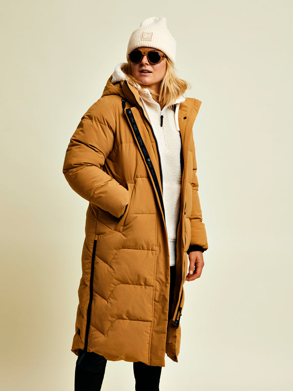 Person, die einen langen braunen Mantel, einen weißen Kapuzenpulli, eine weiße Mütze und eine Sonnenbrille trägt und vor einem hellen Hintergrund steht.