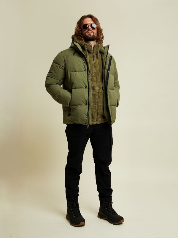 Ein Mann, der eine Sonnenbrille, eine olivgrüne Pufferjacke und eine schwarze Hose trägt, steht vor einem schlichten beigen Hintergrund.