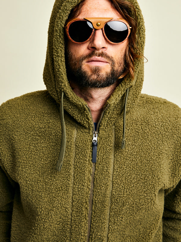 Mann mit olivgrünem Fleece-Kapuzenpulli und runder Holzsonnenbrille, Blick nach vorn vor hellem Hintergrund.