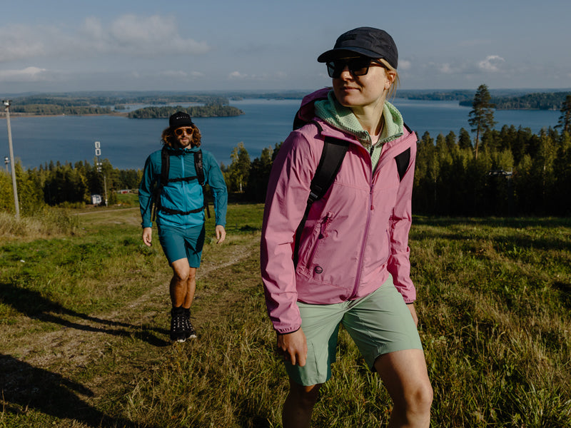 Zwei Personen mit Jacken und Rucksäcken wandern einen grasbewachsenen Hügel hinauf, im Hintergrund sind ein See und Bäume zu sehen.