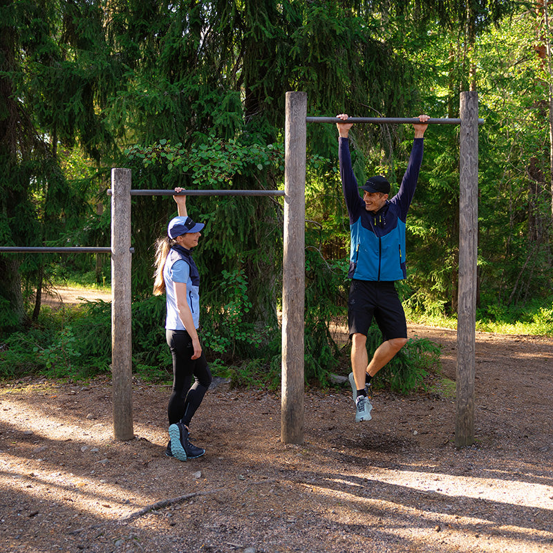 Ein Mann hängt an einer Klimmzugstange, während eine Frau in der Nähe in einem bewaldeten Gebiet mit Fitnessgeräten steht.