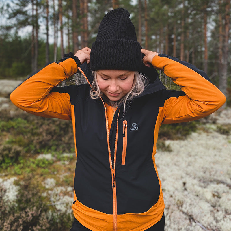 Eine Person in einer Halti Pallas Plus III Damen Warm Hybrid Jacke und einer schwarzen Mütze richtet ihre Kapuze draußen in einem Wald.