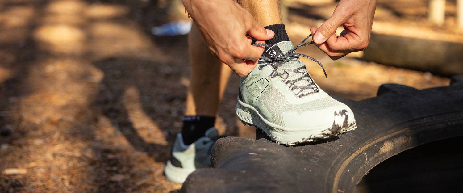 Person, die die Schnürsenkel eines hellgrünen Laufschuhs bindet, während sie im Freien auf einem großen schwarzen Reifen steht.