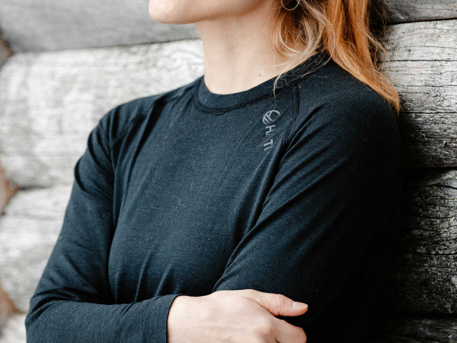 Eine hellhaarige Person in einem schwarzen langärmeligen Hemd steht mit verschränkten Armen vor einer Holzwand.