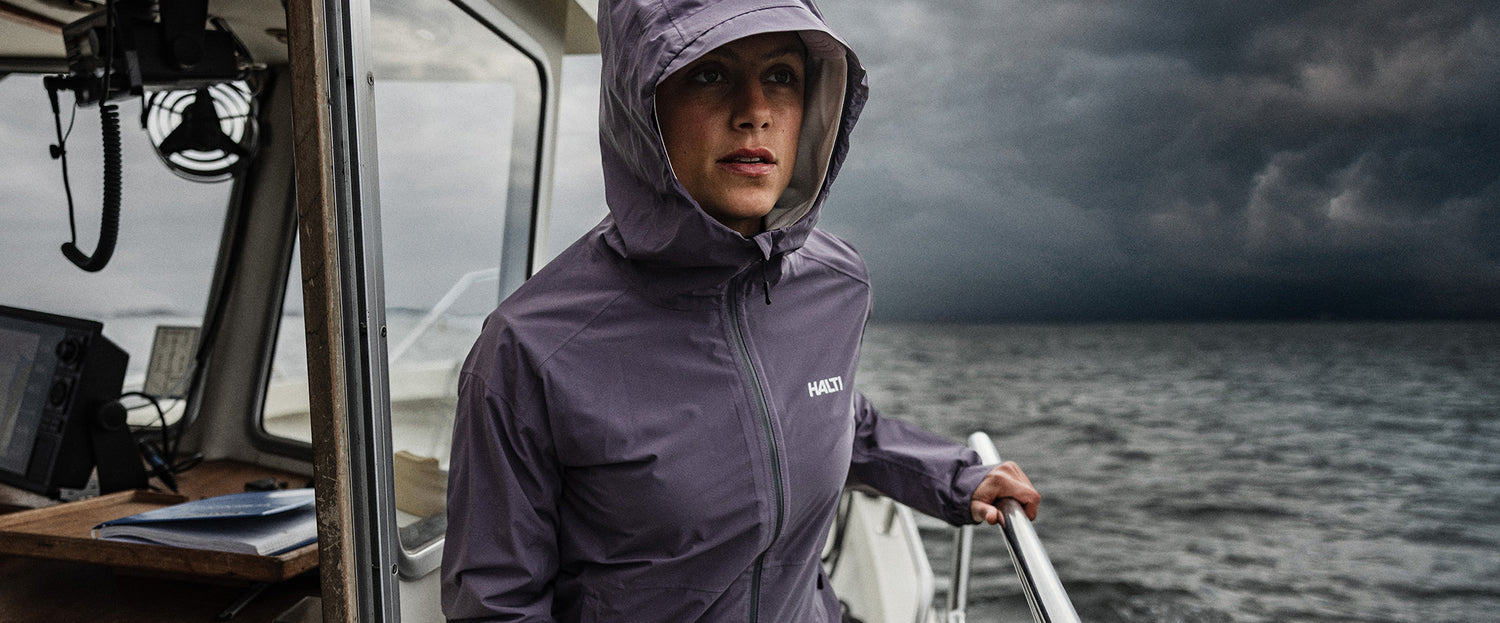 Eine Person in einer lila Regenjacke steht auf einem Boot und hält eine Reling, während hinter ihnen dunkle Gewitterwolken über dem Meer hängen.