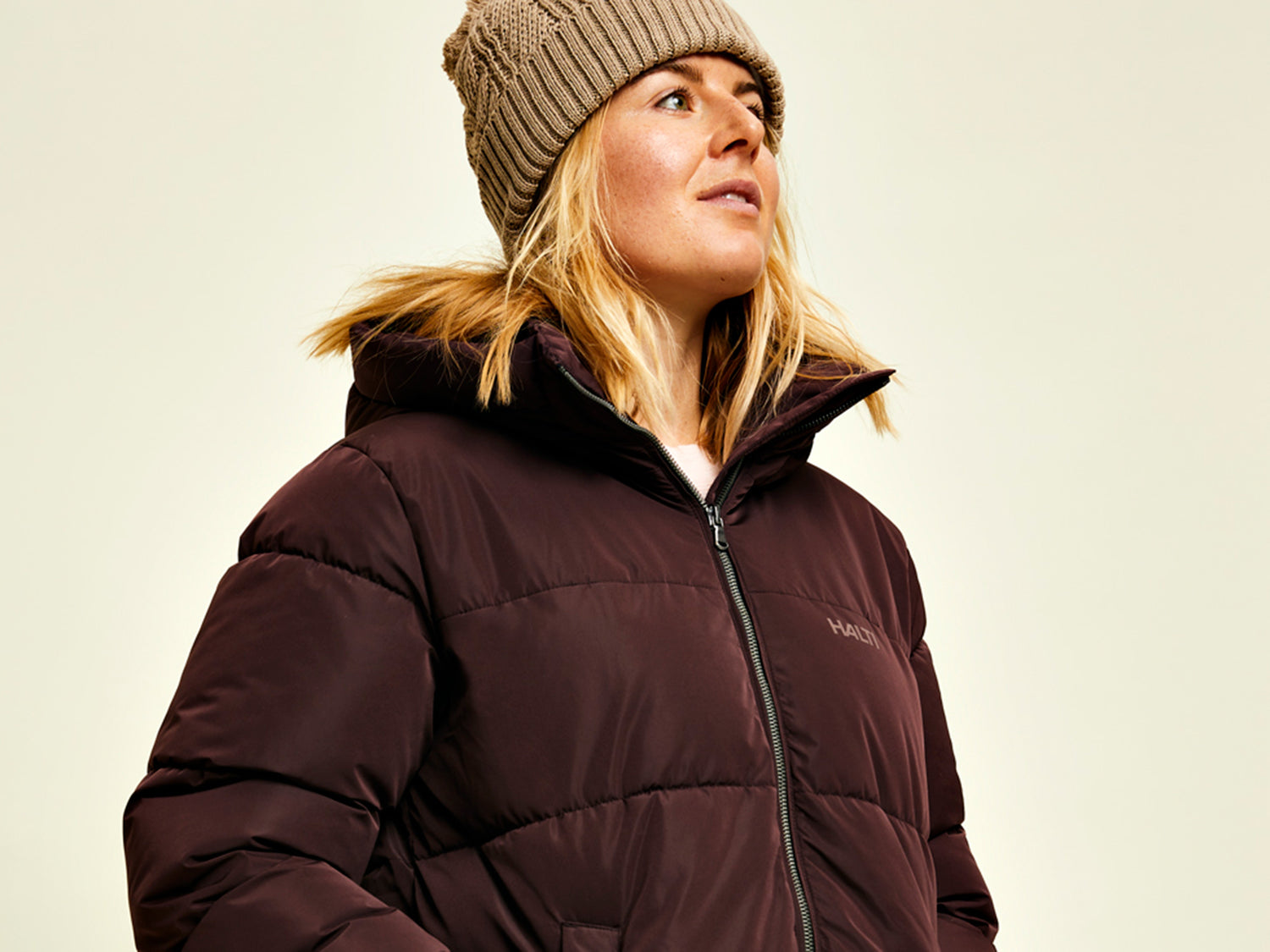 Eine Person in einer braunen Pufferjacke und einer Strickmütze blickt vor einem schlichten Hintergrund leicht nach oben.