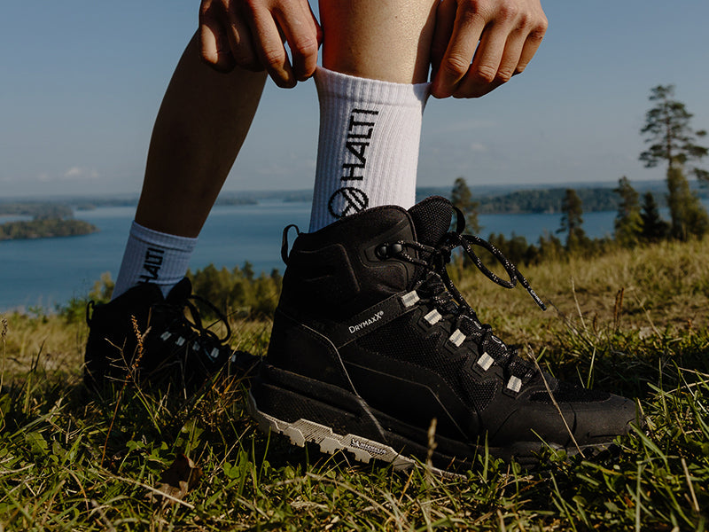 Person auf einer Wiese, die schwarze Wanderschuhe schnürt und weiße Socken mit der Aufschrift HALTI trägt; See und Bäume im Hintergrund.
