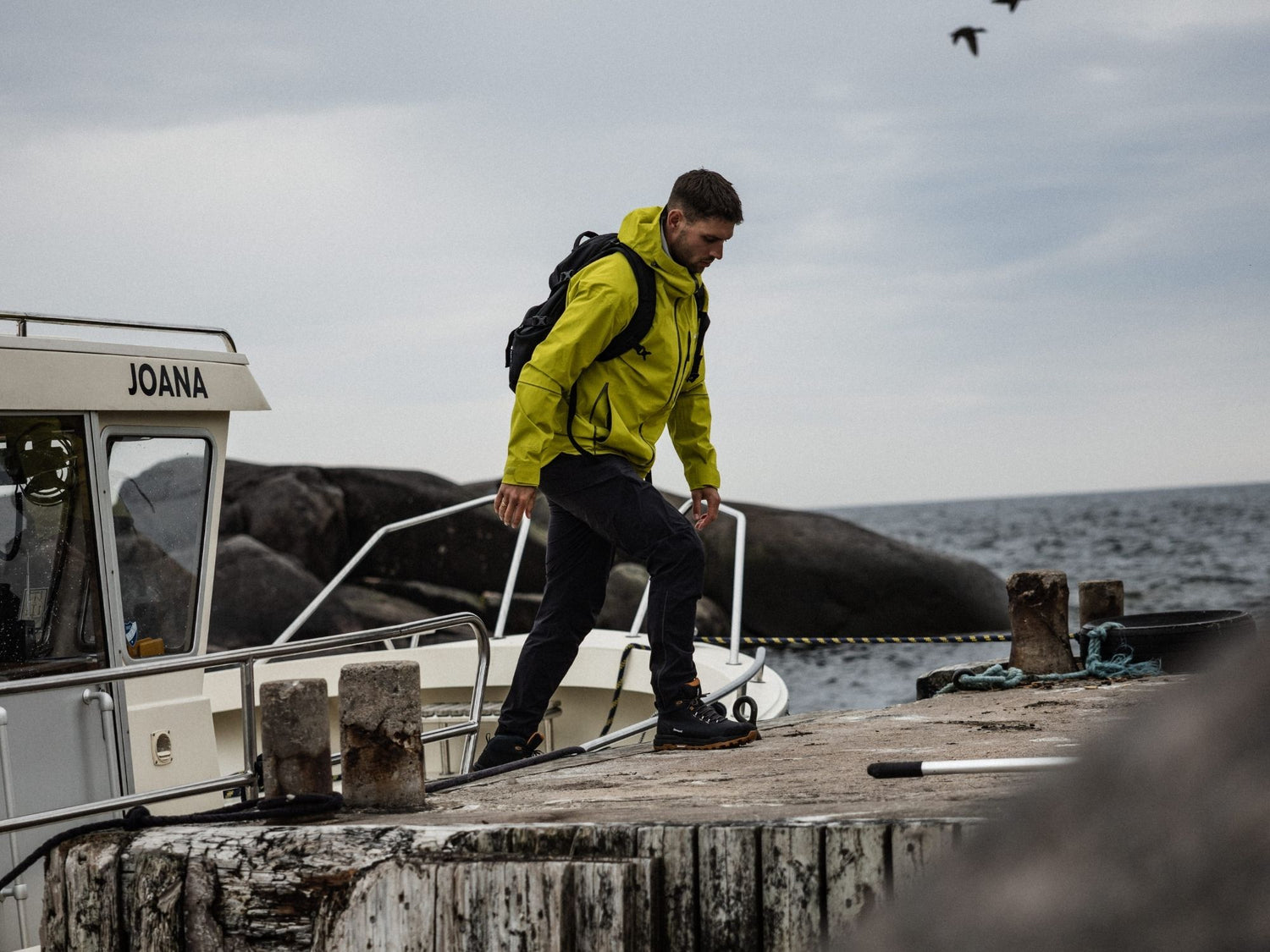 Ein Mann in einer gelben Jacke und mit einem Rucksack steigt von einem Boot auf einen hölzernen Steg am Meer unter einem bewölkten Himmel.