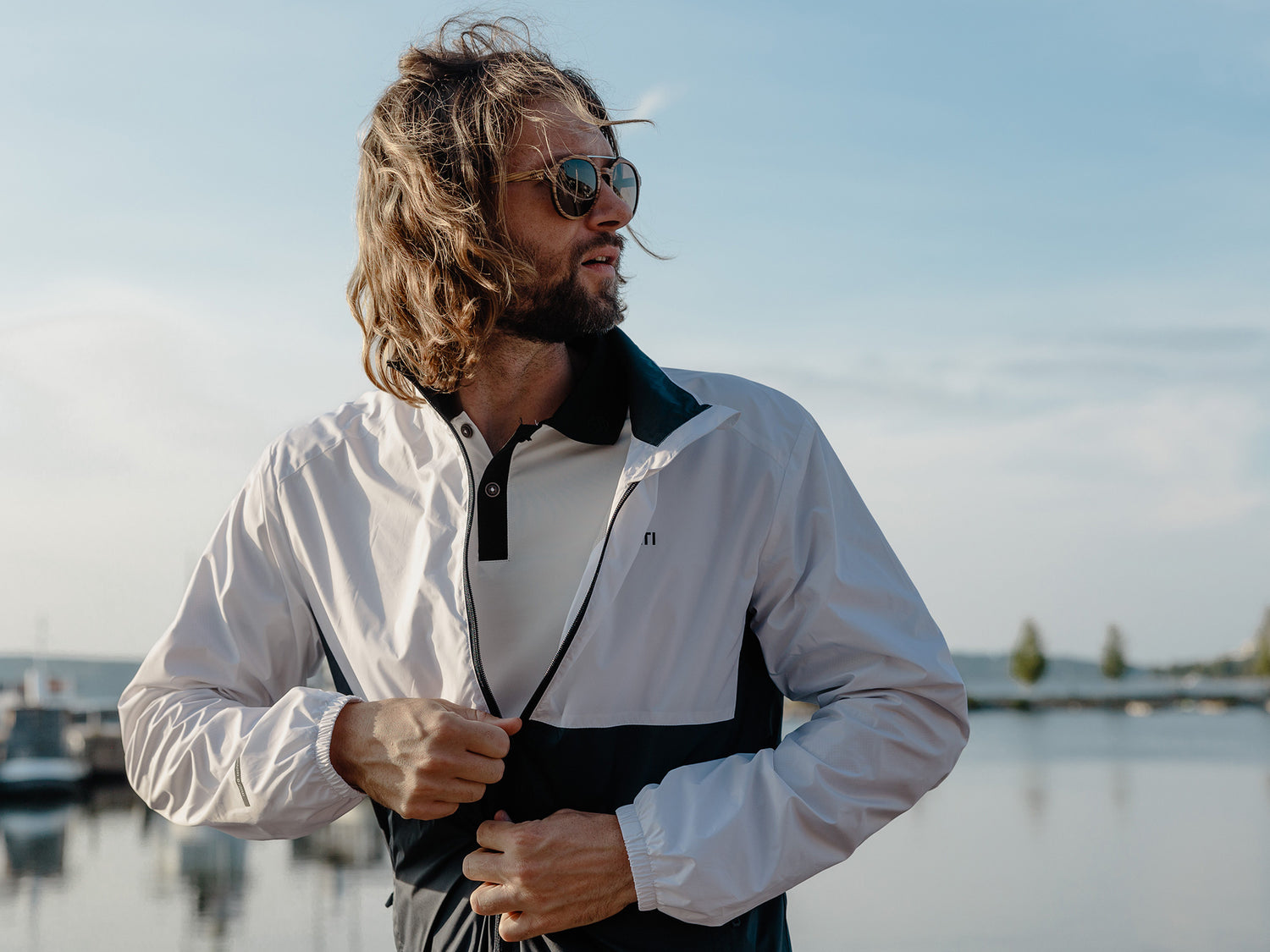 Ein Mann mit langen Haaren und Sonnenbrille schließt den Reißverschluss seiner Jacke, während er in der Nähe eines Gewässers mit Booten im Hintergrund steht.