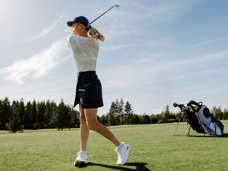 Eine Person mit einer Mütze schwingt einen Golfschläger auf einem Golfplatz mit Bäumen und einer Golftasche im Hintergrund.