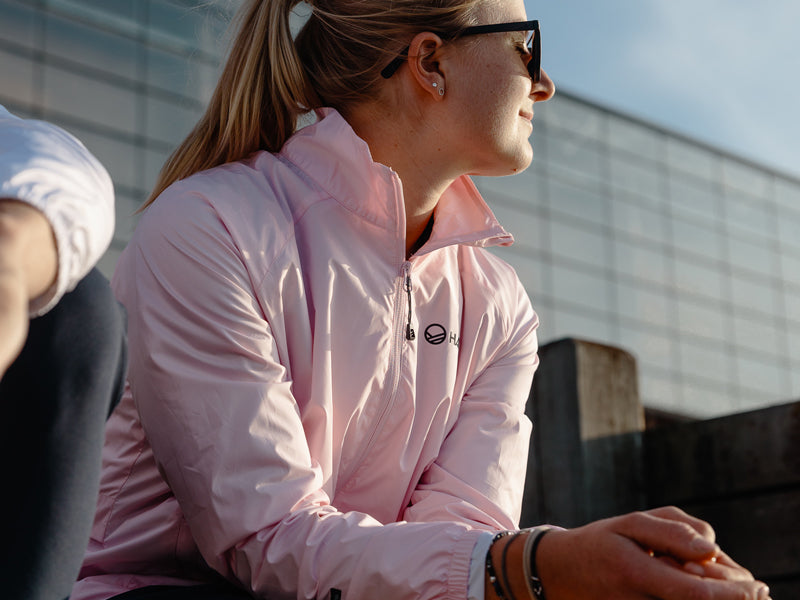 Eine Frau in einer hellrosa Jacke und mit Sonnenbrille sitzt im Freien und blickt zur Seite, mit einem modernen Gebäude im Hintergrund.