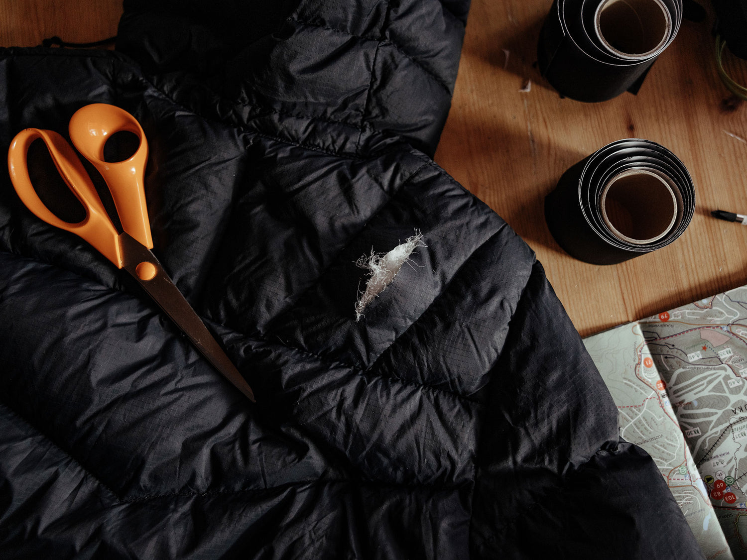 Eine schwarze Pufferjacke mit einem Riss, eine orangefarbene Schere und Reparaturband liegen auf einem Holztisch.