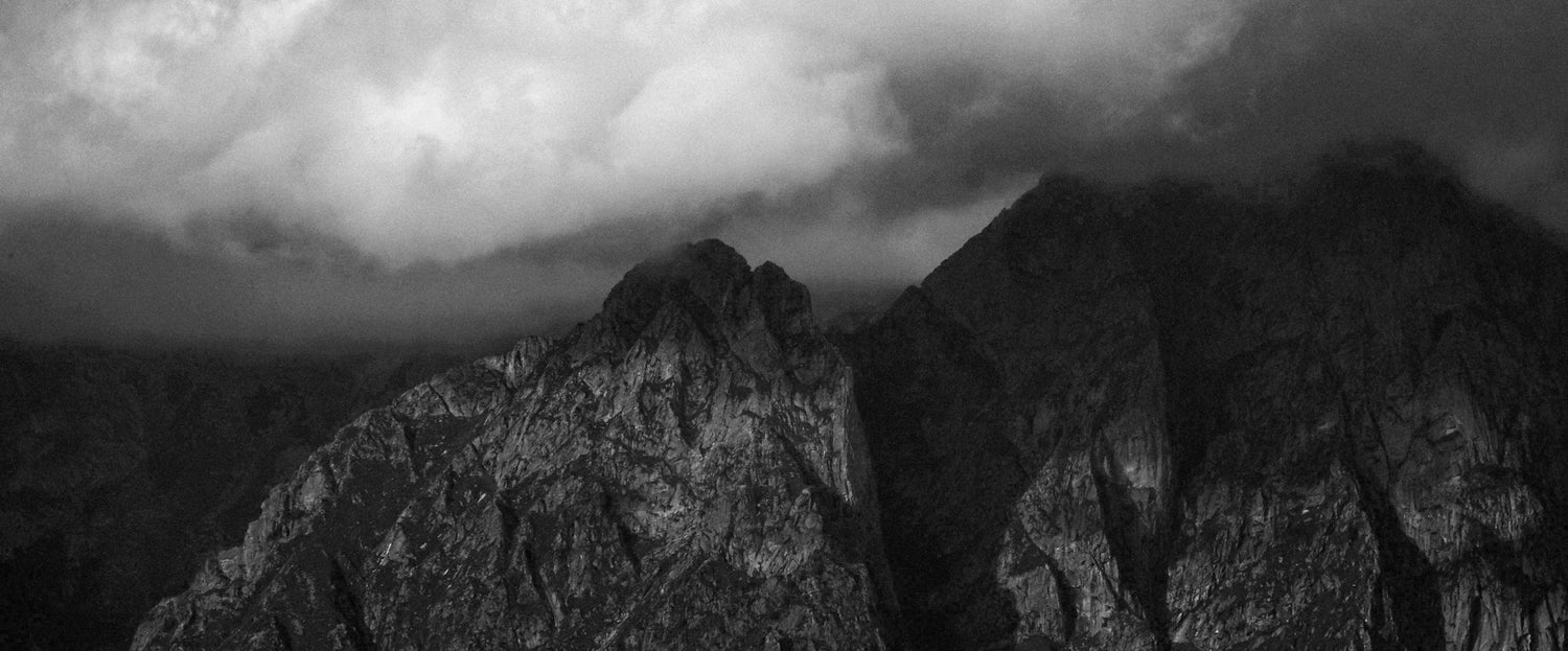 Dunkle, zerklüftete Berggipfel mit schweren Wolken über dem Kopf, die eine dramatische, stimmungsvolle Atmosphäre in Schwarz-Weiß schaffen.