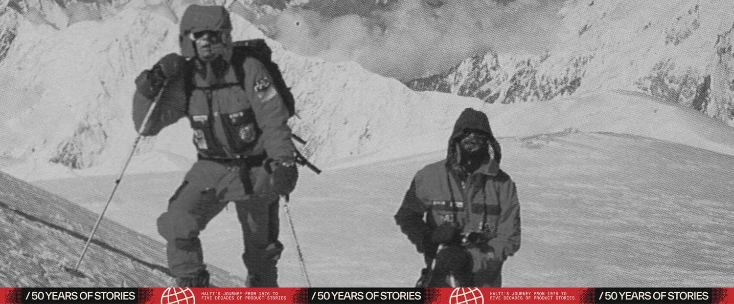 Zwei Bergsteiger in Winterausrüstung stehen an einem verschneiten Berghang mit schneebedeckten Gipfeln im Hintergrund.