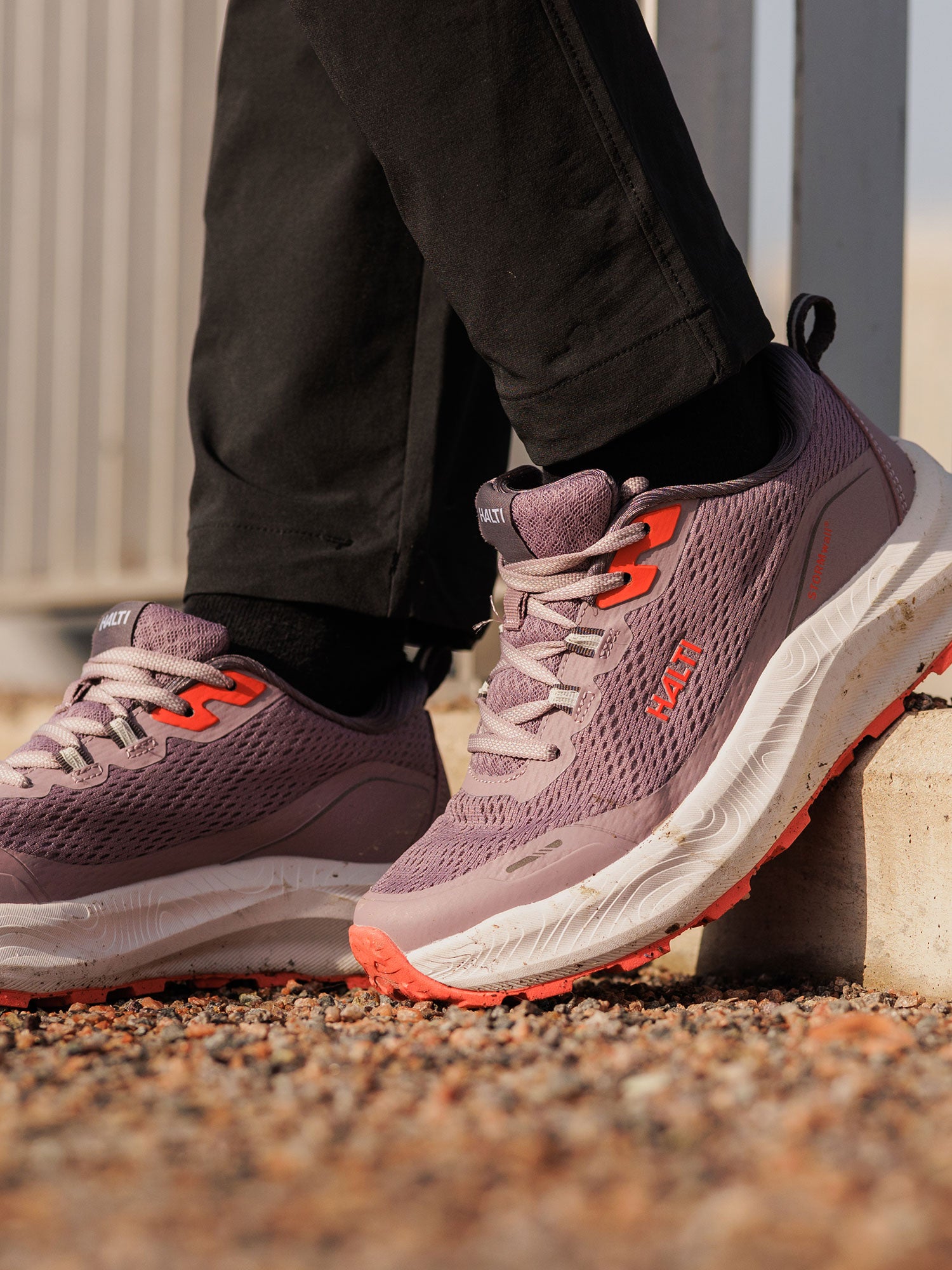 Eine Person in Halti Rapid 2 Sneaker Damen, lila/orange, steht in schwarzen Hosen auf Kies; die Schuhe sind wasserabweisend.