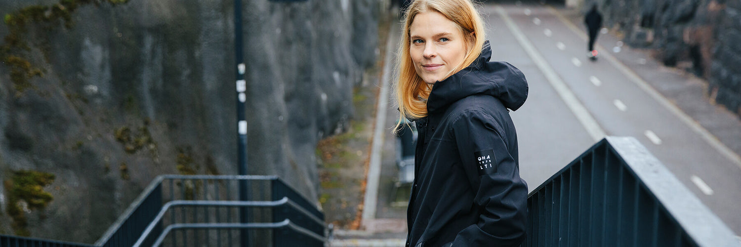 Eine Frau in einer schwarzen Jacke steht auf einer Außentreppe neben einer Felswand und schaut in die Kamera.