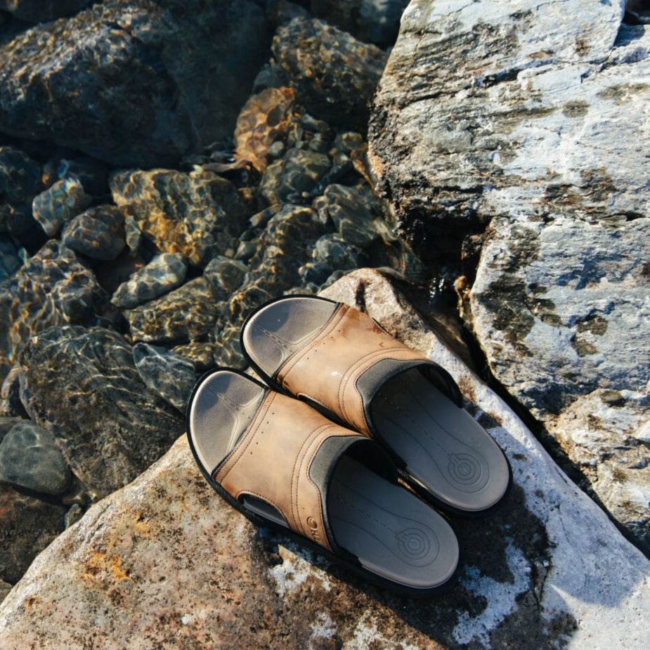 Ein Paar Halti Jorah Herren Sandalen liegt auf einem Felsen am klaren Wasser mit sichtbaren Steinen.