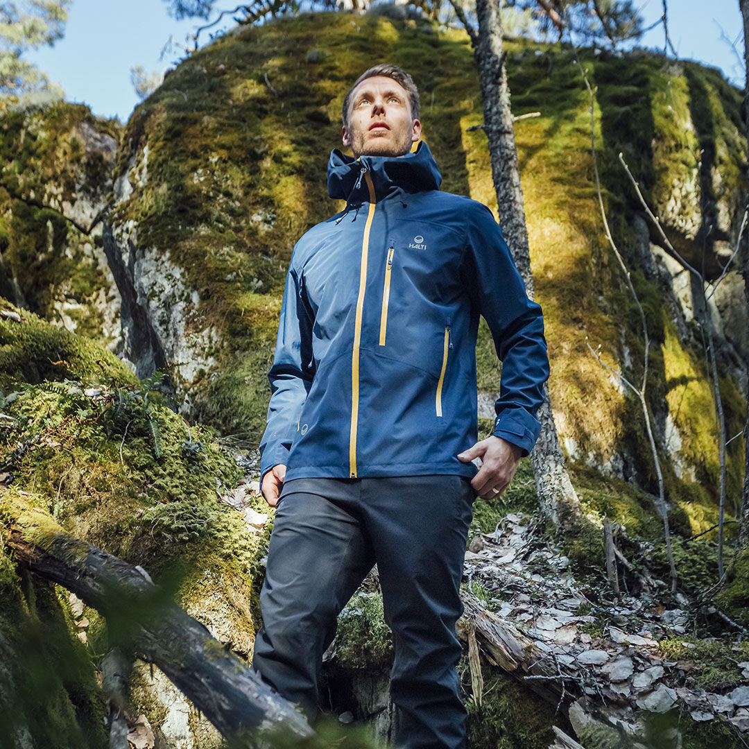 Ein Mann in einer blauen Jacke und Halti Vuoksi DX Regenhose Herren steht in einem Wald, in dem die Sonne auf moosbewachsene Felsen scheint.