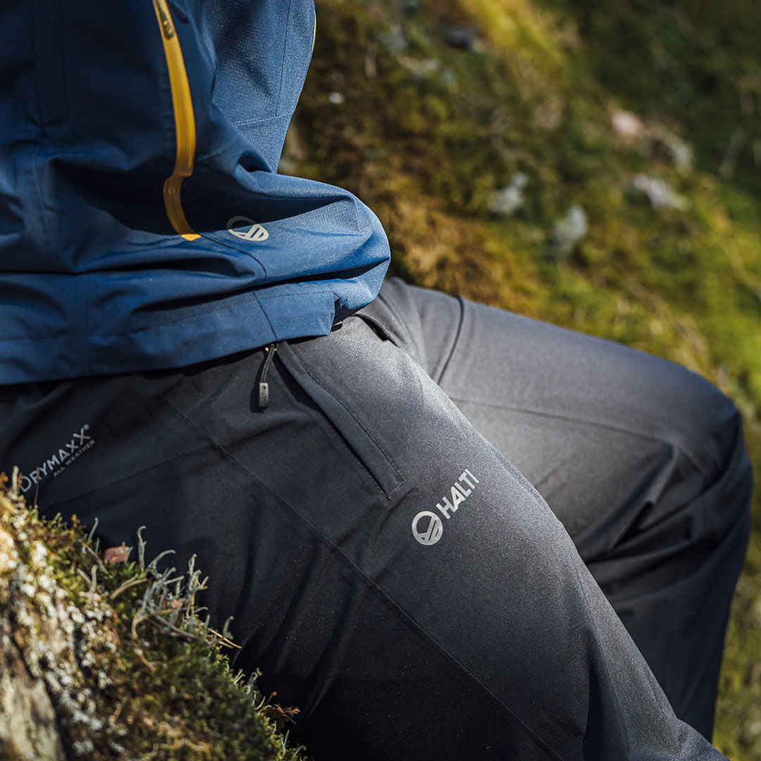 Person in Halti Vuoksi DX Regenhose sitzt draußen auf moosigem Boden, trägt blaue Jacke mit gelbem Reißverschluss.