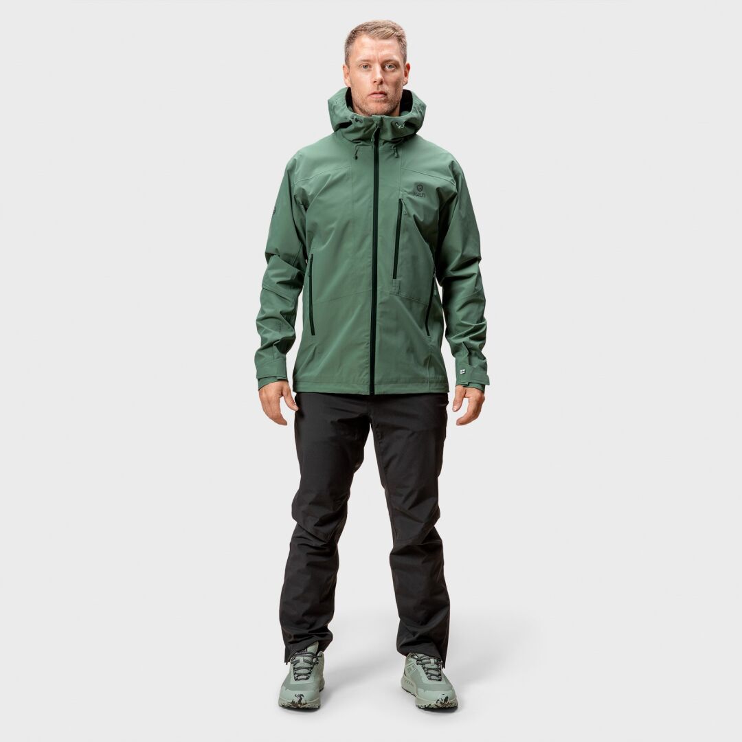 Ein Mann trägt die Halti Juonto DrymaxX Regenjacke für Herren sowie schwarze Hose und grüne Schuhe.