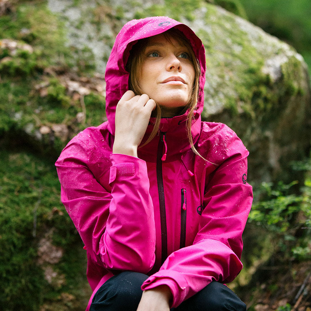 Jemand in einer leuchtend rosa Halti Biegga DX Regenjacke Damen sitzt im Freien mit Grünzeug und Steinen im Hintergrund.