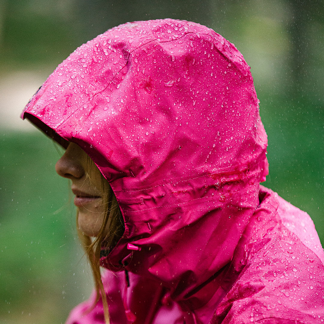 Jemand trägt eine leuchtend rosa Halti Biegga DX Regenjacke Damen mit aufgesetzter Kapuze, auf der Regentropfen abperlen.