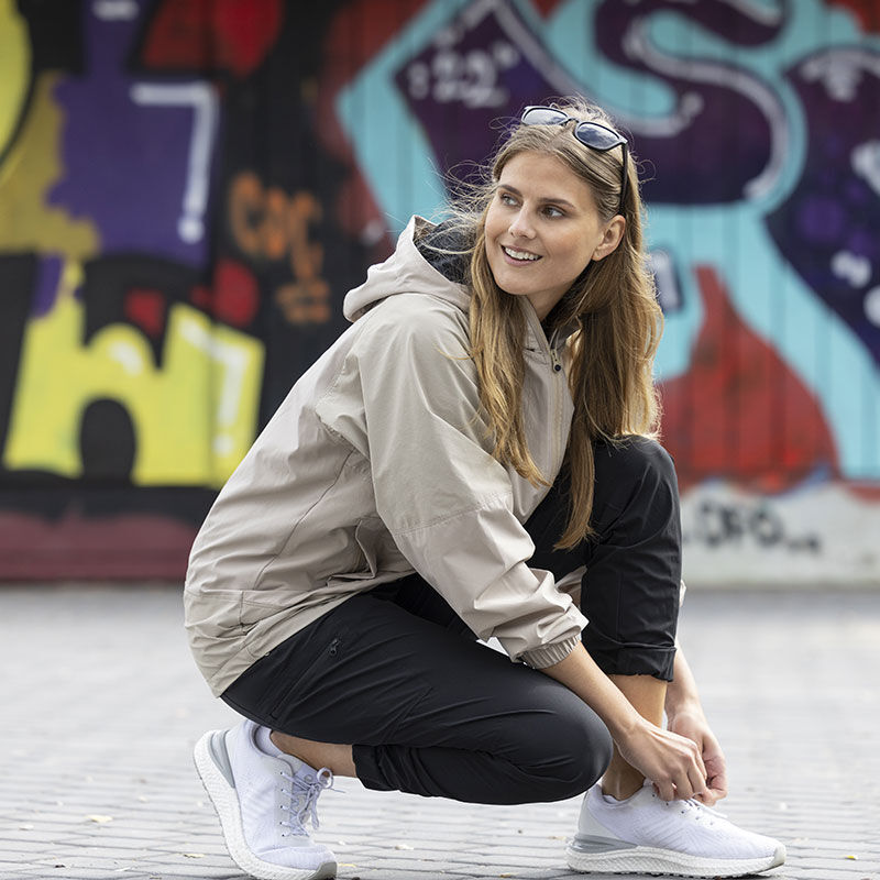 Frau in beiger Jacke und schwarzer Hose bindet ihren Halti Gale Sneaker Damen mit Obermaterial aus recyceltem Textil an einer Graffiti-Wand fest.
