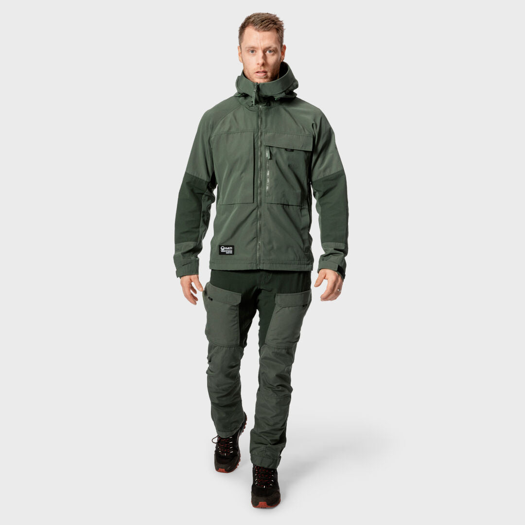 Ein Mann in einer dunkelgrünen, atmungsaktiven Outdoorjacke und einer belüfteten Halti Hiker Herren-Hose mit schwarzen Stiefeln steht vor einem grauen Hintergrund.