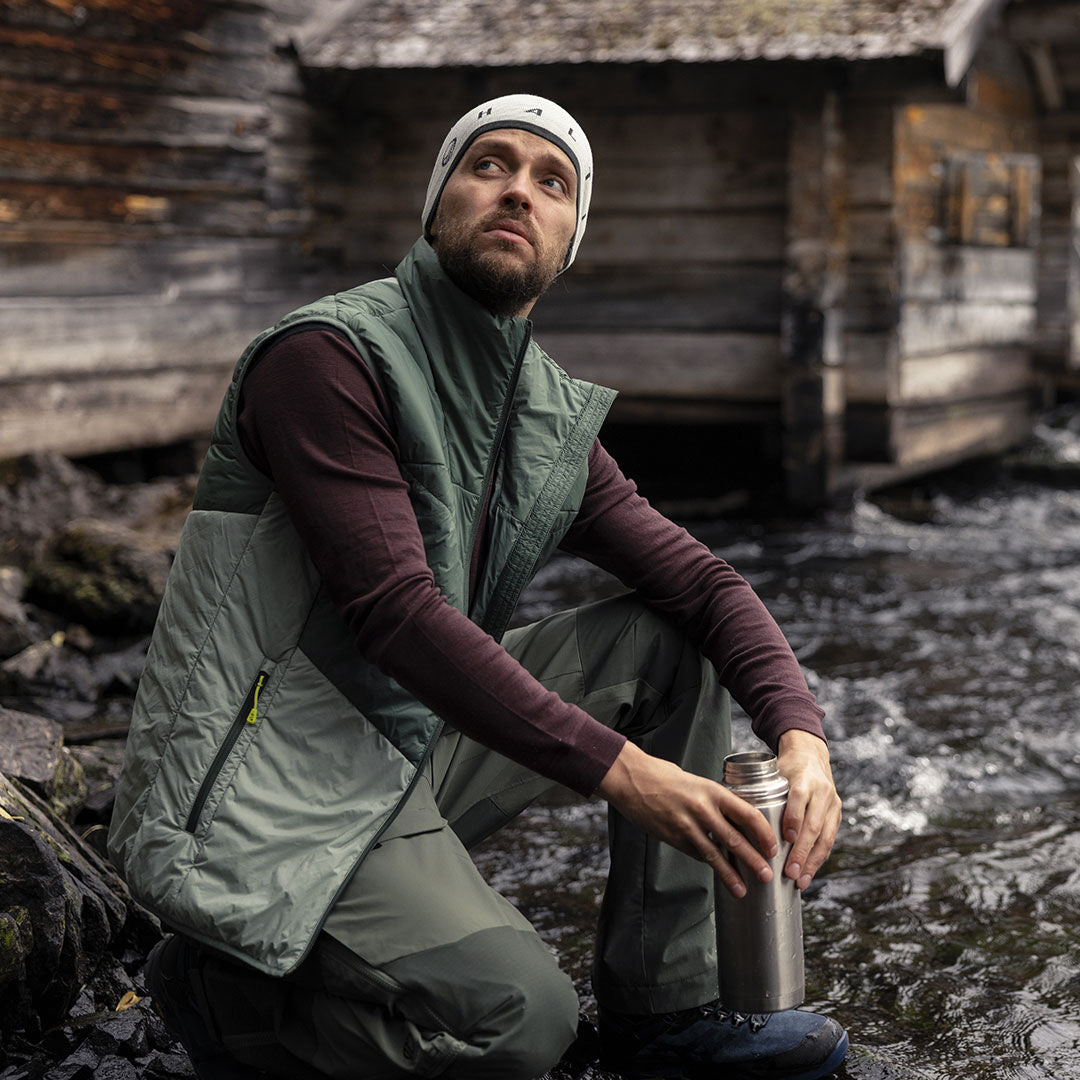 Ein Mann in einer Halti Heatix Primaloft Weste Herren kniet mit einer Metallflasche an einem Bach; hinter ihm befinden sich Holzhütten.