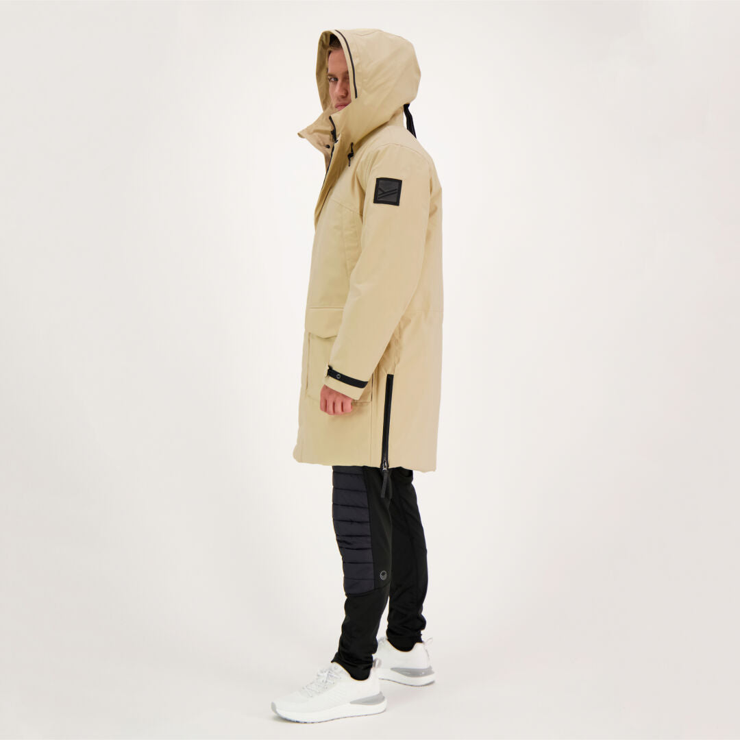 Eine Person in einem beigen Halti Bergga DX Winterparka, einer schwarzen Hose und weißen Turnschuhen steht seitlich auf einem weißen Hintergrund.