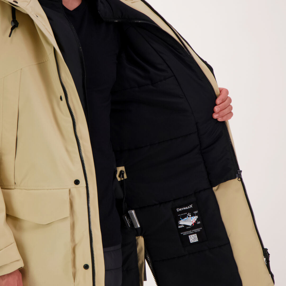Ein Mann öffnet einen beigefarbenen Halti Bergga DX Winterparka und enthüllt das schwarze Isolierfutter und das aufgenähte Etikett.