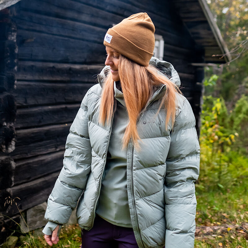 Eine Person in einer Halti Versant Daunenjacke für Damen und einer braunen Mütze steht draußen bei einer Hütte im Grünen.