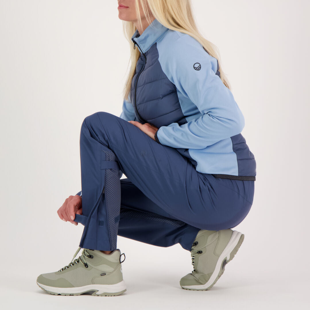 Eine Frau mit langen blonden Haaren hockt in einer blauen Halti Liike Damen 3L DrymaxX Sporthose und hellgrauen Wanderstiefeln.