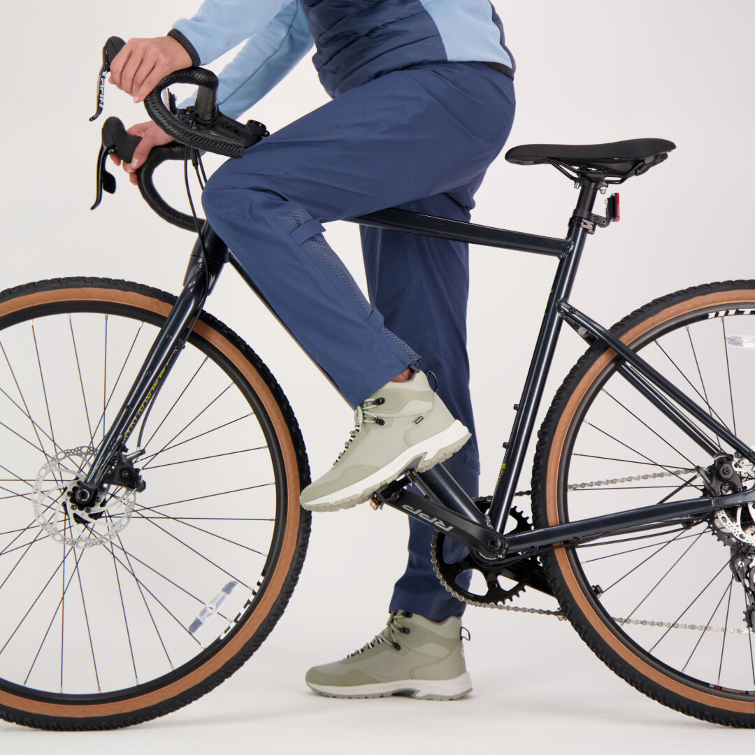 Frau in blauer Halti Liike Damen 3L DrymaxX Sporthose und grauen Turnschuhen fährt auf einem schwarzen Fahrrad auf weißem Hintergrund.