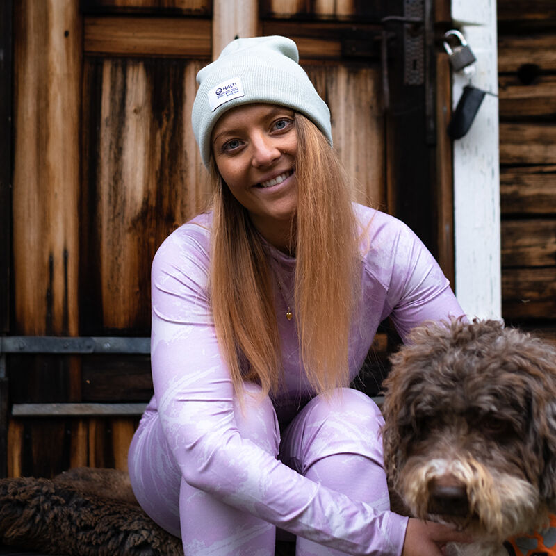 Eine Frau im Halti Windfire Baselayer Set Damen und einer grauen Mütze sitzt auf Stufen und streichelt ihren braunen, lockigen Hund.