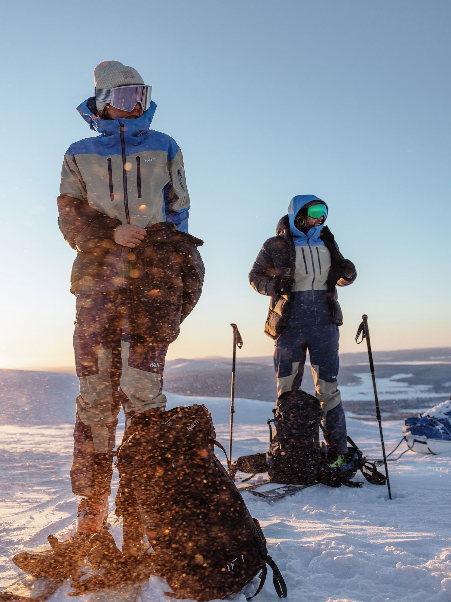 Zwei Personen in Winterjacken, Hosen, Mützen und Schutzbrillen stehen in einer verschneiten Landschaft mit Rucksäcken und Trekkingstöcken, in der Sonne und Schneepartikel in der Luft liegen.