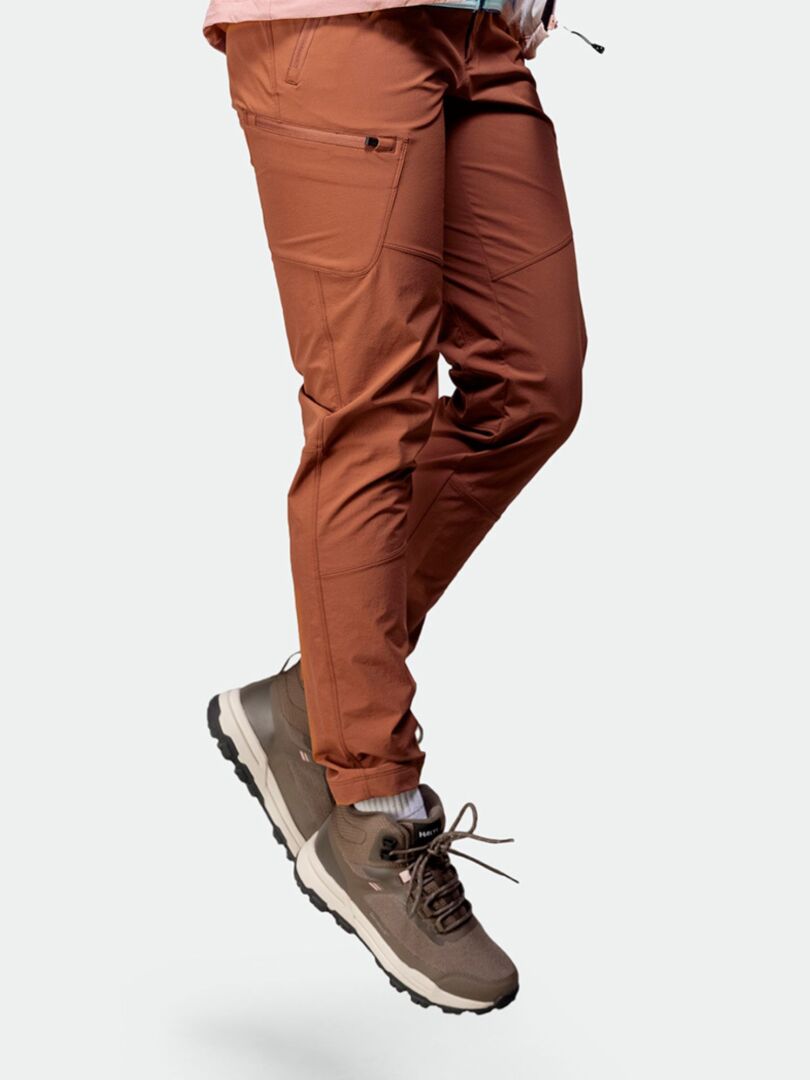 Person in brauner Outdoor-Hose und Halti Fara 3 DX Mid Outdoor-Schuhen Damen, dargestellt mit der Taille nach unten vor einem schlichten Hintergrund.