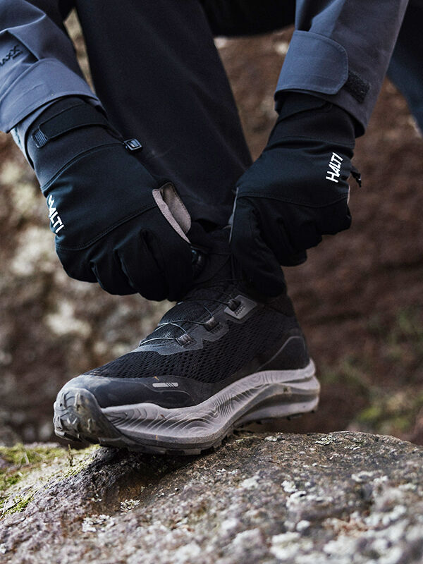 Eine Person mit schwarzen Handschuhen bindet das Freelock-System von Halti Rapid Mid Freelock DX Schuhe Unisex an einen Felsen im Freien.