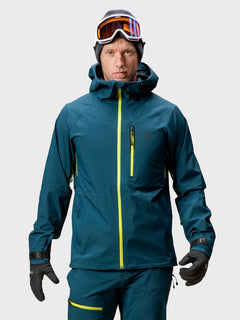Eine Person in blauer Skikleidung trägt die Halti Alpine 3L DX Skijacke Unisex, bereit zum Skifahren im Hinterland.