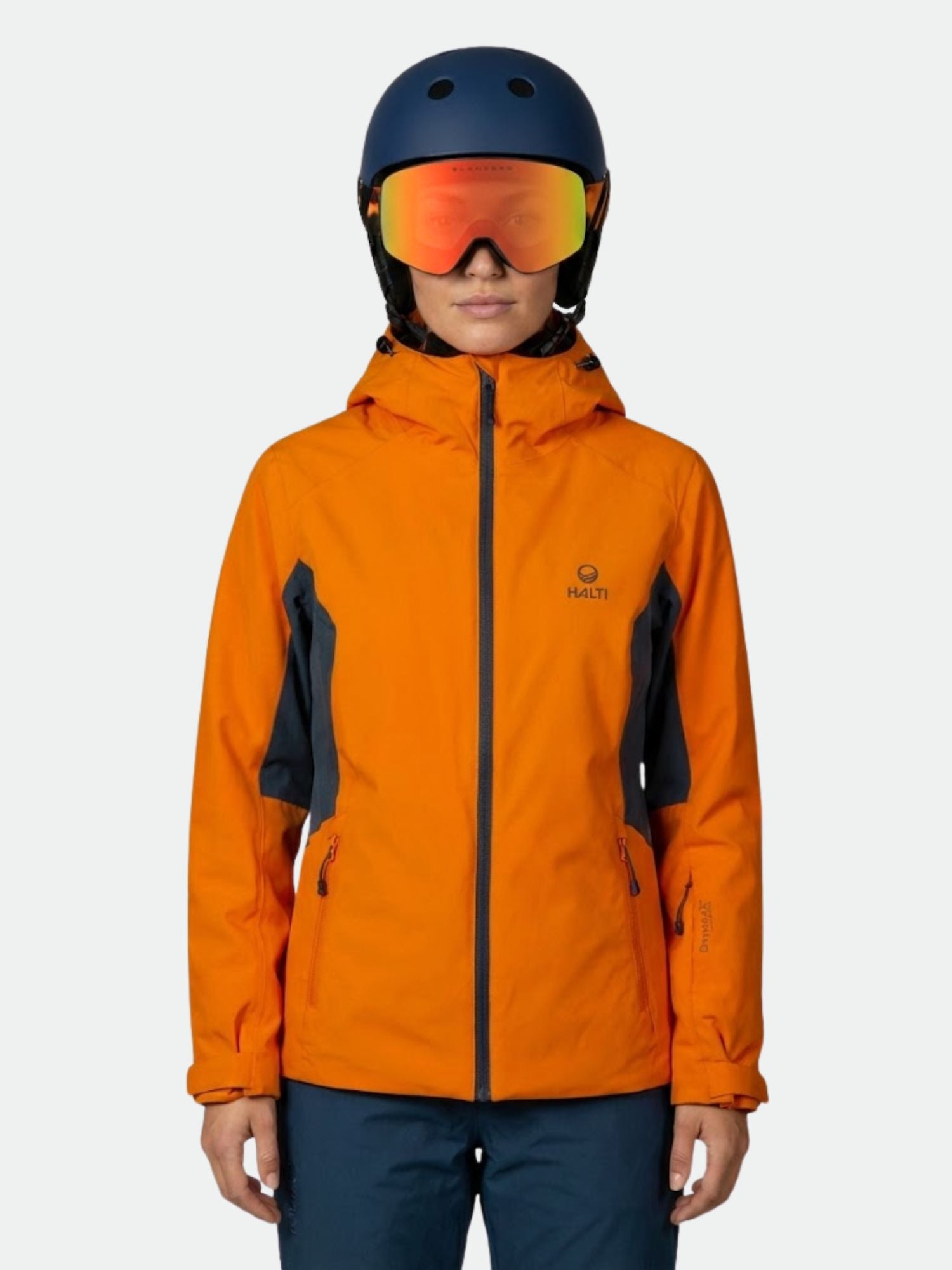 Eine Person in einer Halti Tahko DX Skijacke Damen, einer blauen Hose, einem Helm und einer Skibrille steht vor einem schlichten Hintergrund.