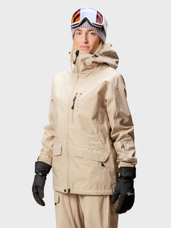 Eine Person in einer beigen Halti Settler 3L DrymaxX Ski Jacke Damen, beiger Hose und Skiausrüstung steht vor einem schlichten Hintergrund.