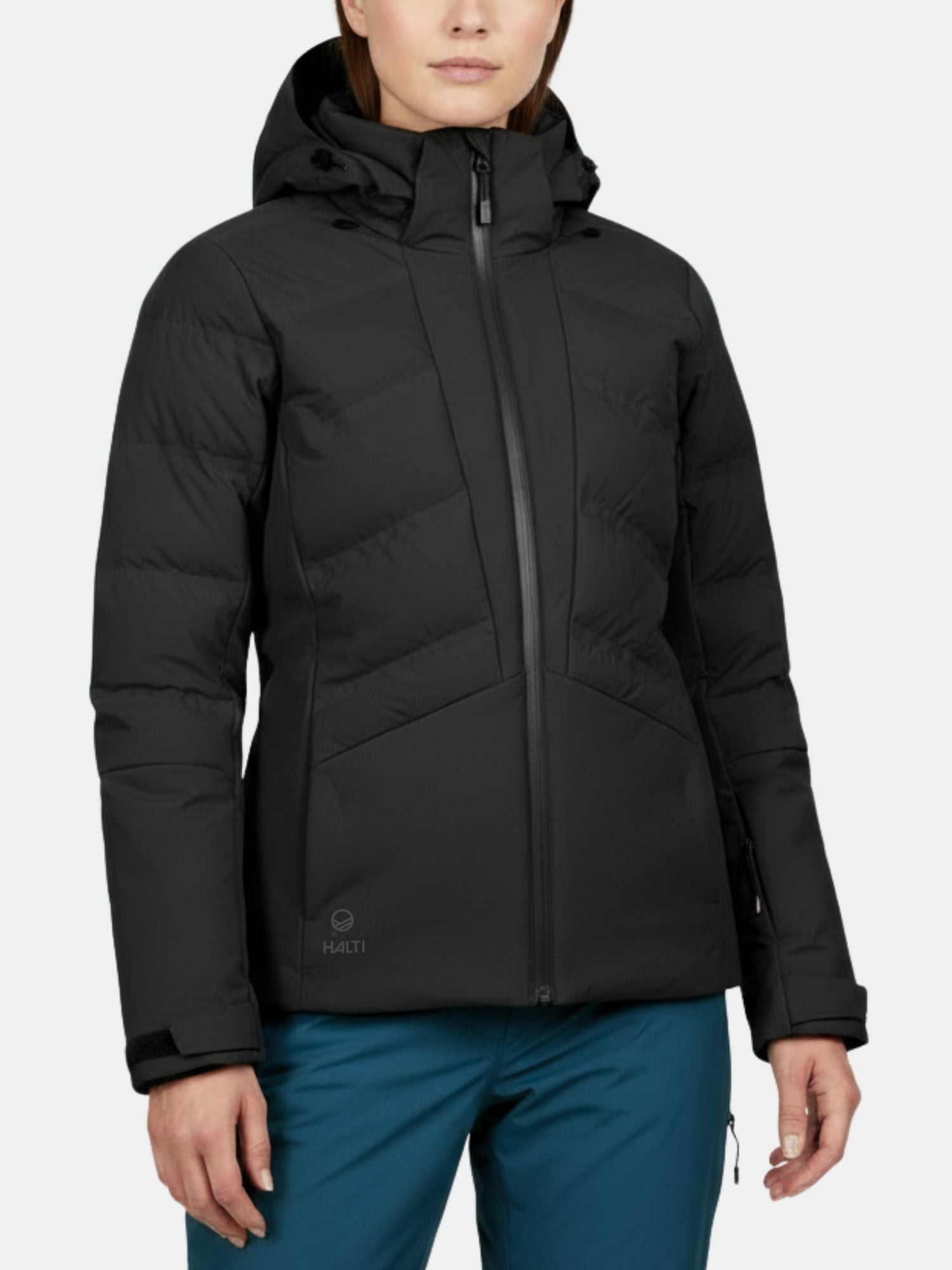 Eine Person in einer schwarzen Halti Nordic Arcty Skijacke Damen mit Frontreißverschluss und blauer Hose, vor einem schlichten Hintergrund.